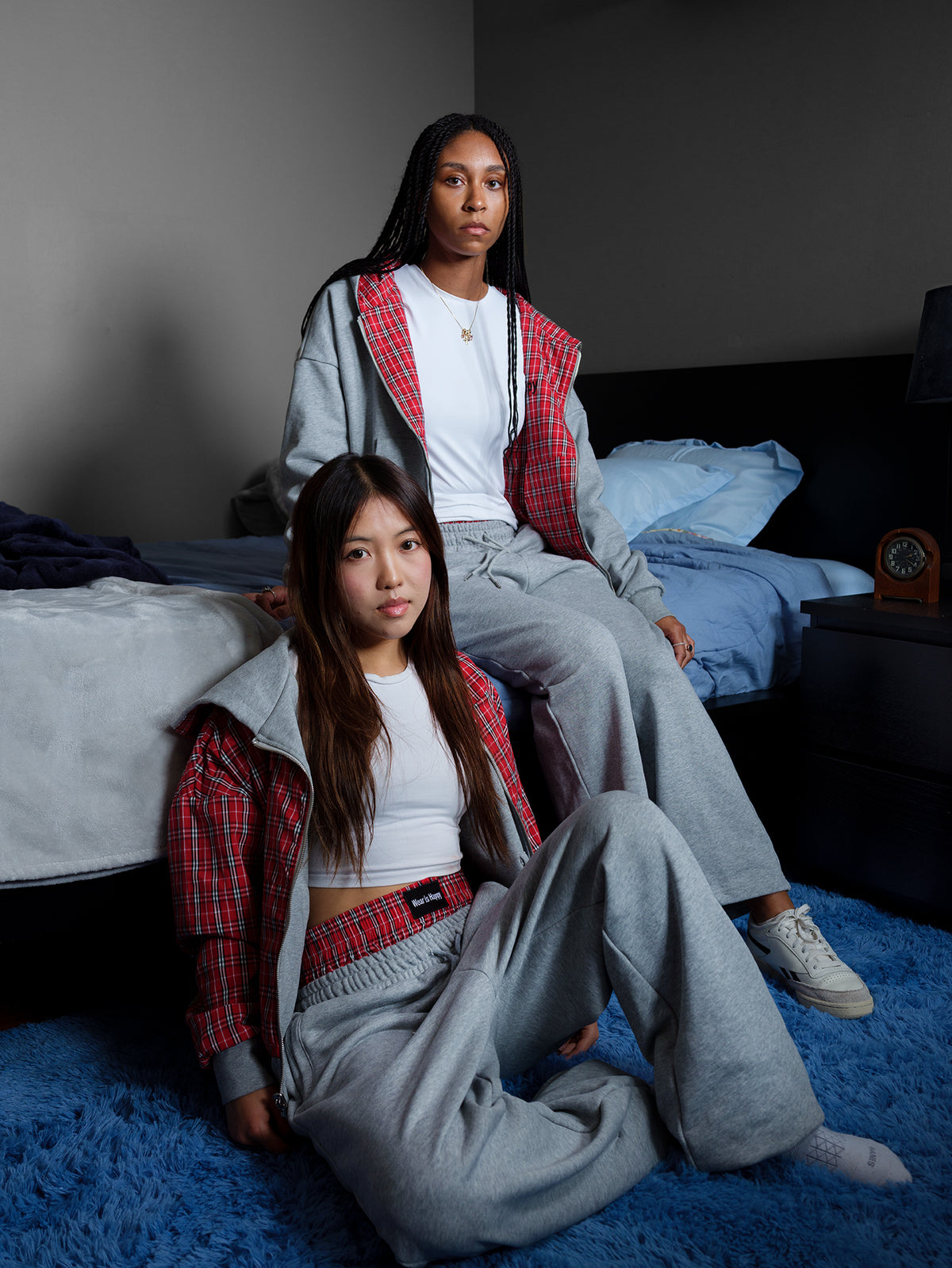 Two women posing in matching gray sweatpants and white crop tops, wearing red and black plaid zip-up overshirts, in a dimly lit bedroom setting.