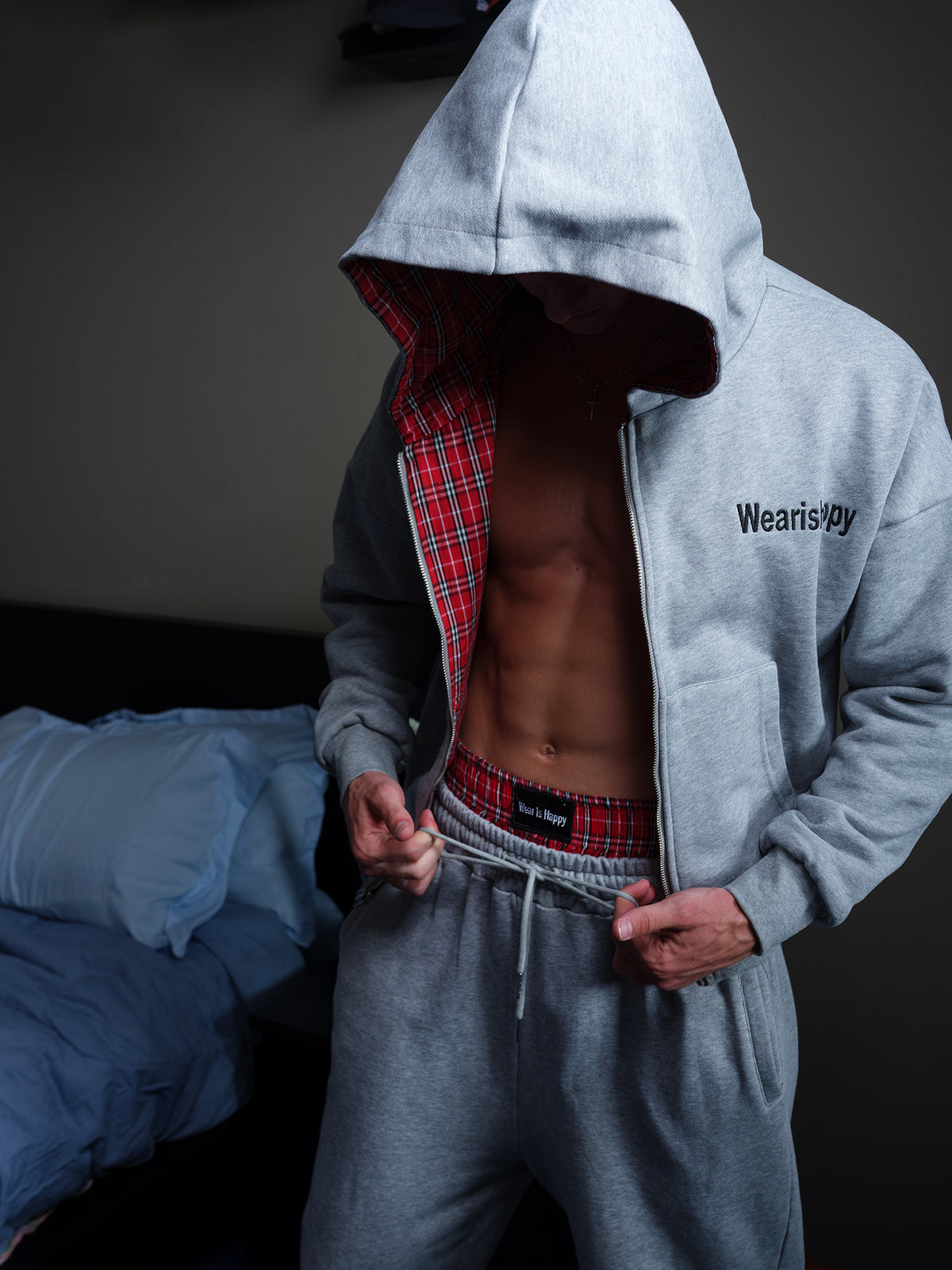 A man wearing a light gray zip-up hoodie with a red plaid lining and matching gray sweatpants, standing in a dimly lit room with a bed in the background.