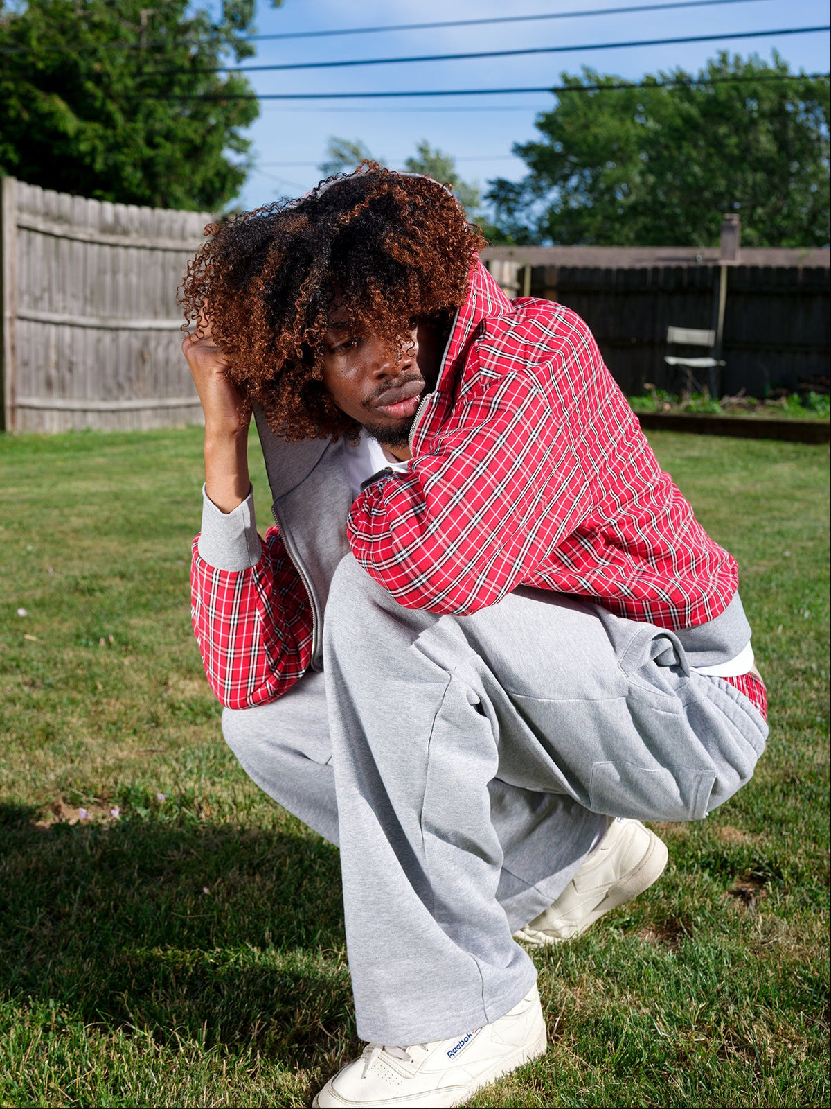 A person crouching outdoors on grass in front of a wooden fence, wearing a red and white plaid button-up shirt layered over a light gray zip-up hoodie, loose gray sweatpants, and white sneakers.