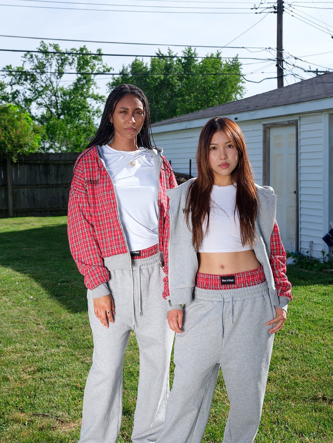 Two women standing outdoors on a grassy lawn in front of a white garage house, wearing matching gray sweatpants, white crop tops, and red-and-white patterned jackets.