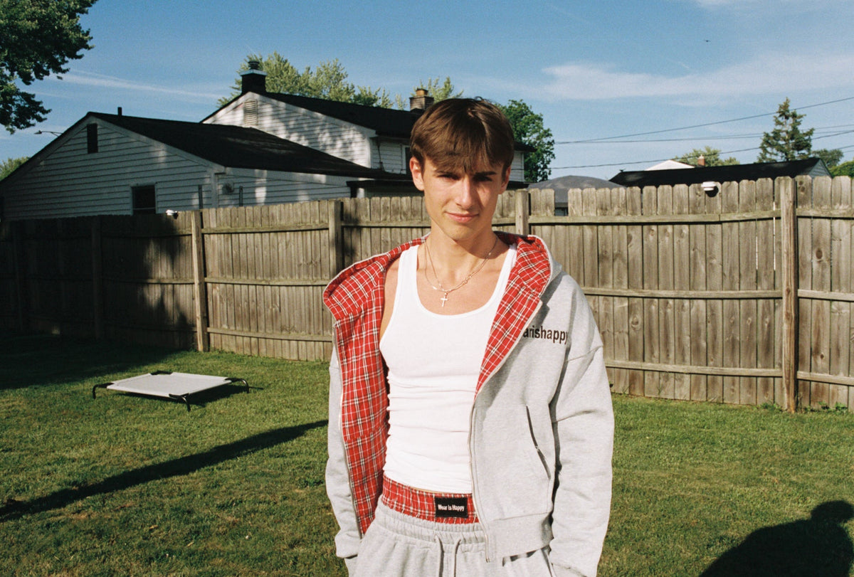 A young man standing in a backyard wearing a light gray zip-up hoodie over a red plaid shirt, a white tank top, and gray sweatpants. A wooden fence, grass, and houses are visible in the background.