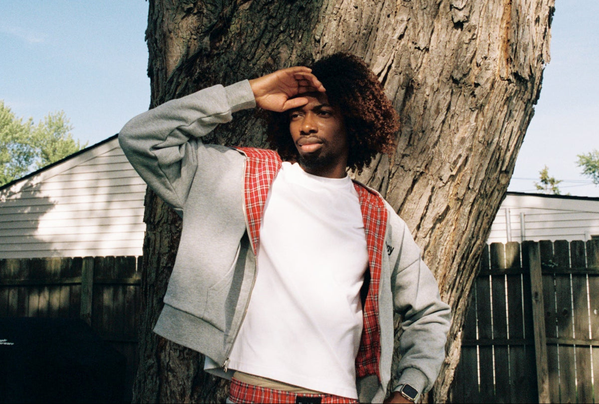 A man with curly hair stands outdoors leaning against a large tree trunk, wearing a light gray zip-up hoodie over a white t-shirt and red-and-white plaid pants.