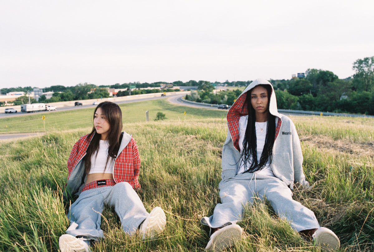 Two women sitting in a grassy field outdoors, wearing streetwear. One wears a red and white plaid flannel shirt over a white crop top and light blue jeans, while the other wears a light gray hoodie with a red-and-white plaid lining and matching sweatpants.